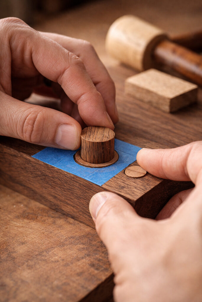 Hands align plug grain with the board before tapping it into a counterbored hole, blue tape guarding the surface from glue.